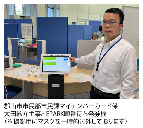 郡山市市民部市民課マイナンバーカード係太田絋介主事とEPARK順番待ち発券機（※撮影用にマスクを一時的に外しております）